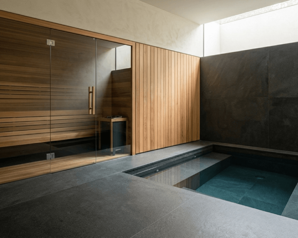 A minimalist, high-end residential contrast therapy circuit. Features include a bespoke Canadian Hemlock infrared sauna with a glass front, sitting adjacent to a recessed cold plunge pool integrated flush into a continuous slab of dark matte basalt stone. Minimalist architecture and soft natural lighting.