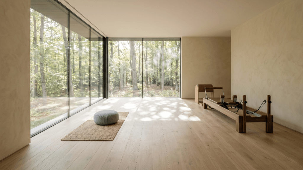 Wide-plank white oak flooring in a modern luxury mind-body studio featuring floor-to-ceiling glass windows overlooking a forest, complete with a bespoke wooden Pilates reformer and a minimalist meditation space.