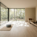 Wide-plank white oak flooring in a modern luxury mind-body studio featuring floor-to-ceiling glass windows overlooking a forest, complete with a bespoke wooden Pilates reformer and a minimalist meditation space.