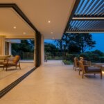 A luxury modern home featuring completely open glass walls and zero-clearance flooring transitioning seamlessly to an outdoor living space with a pergola.